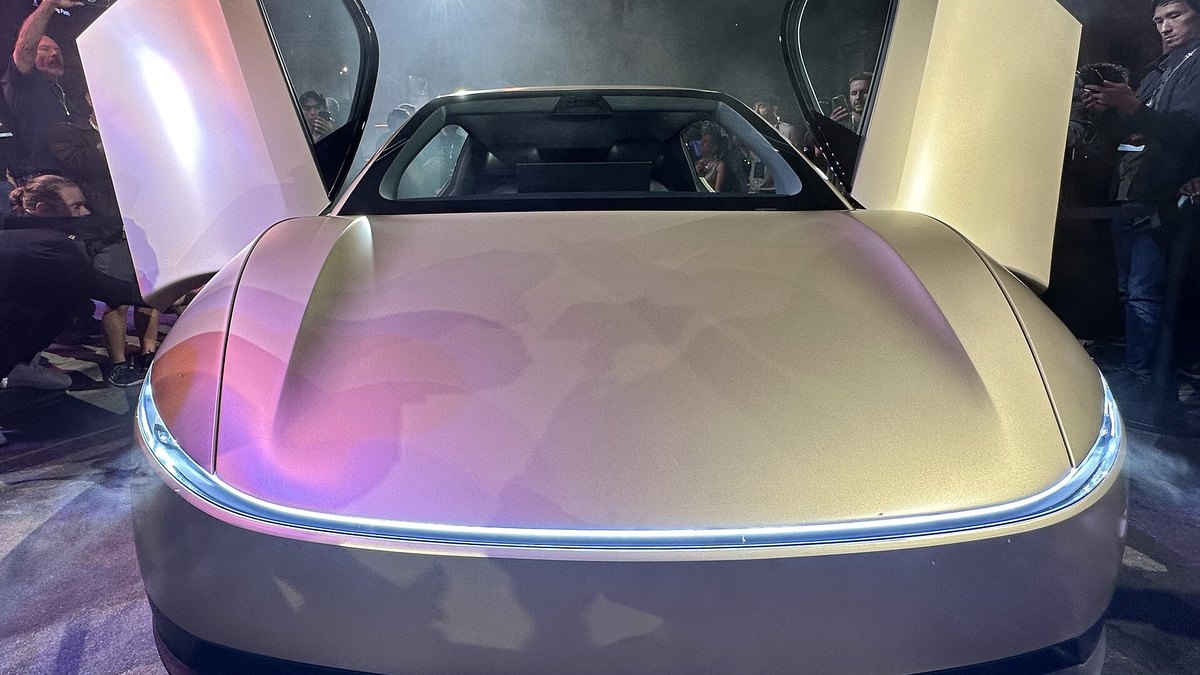 The front of a gold Tesla Cybercab with both doors raised and a light bar across the nose, shown at an unveiling event