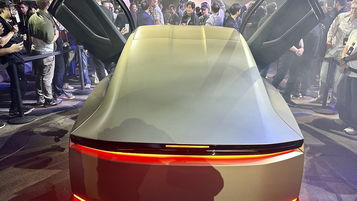 The rear of a gold Tesla Cybercab with a full-width red light bar and doors raised, surrounded by onlookers