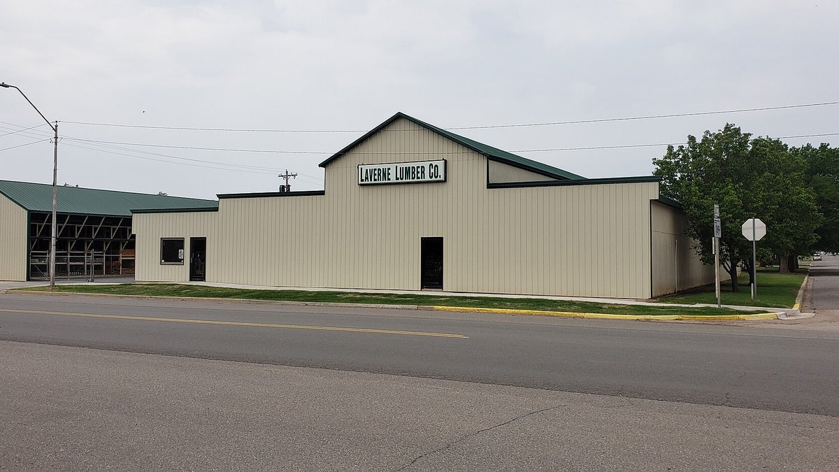 A tan metal-sided Laverne Lumber Co. building with a green roof and open lumber sheds beside a quiet street in Laverne, Oklahoma