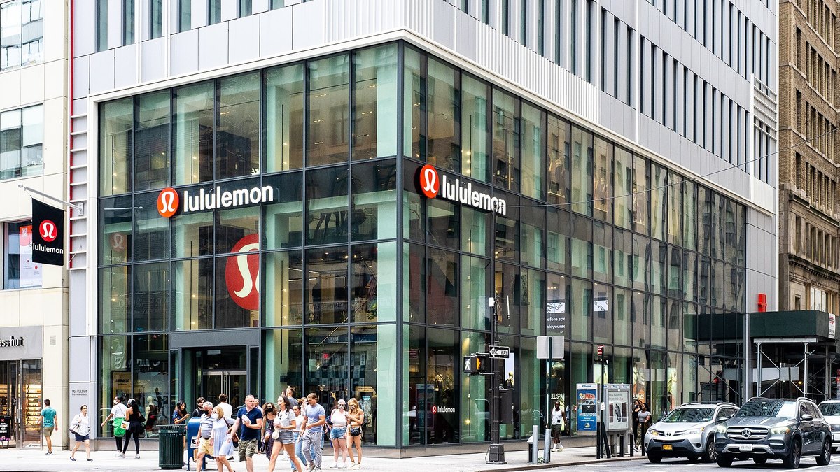 A lululemon store with glass walls and a corner entrance on a busy Manhattan street with pedestrians crossing in front