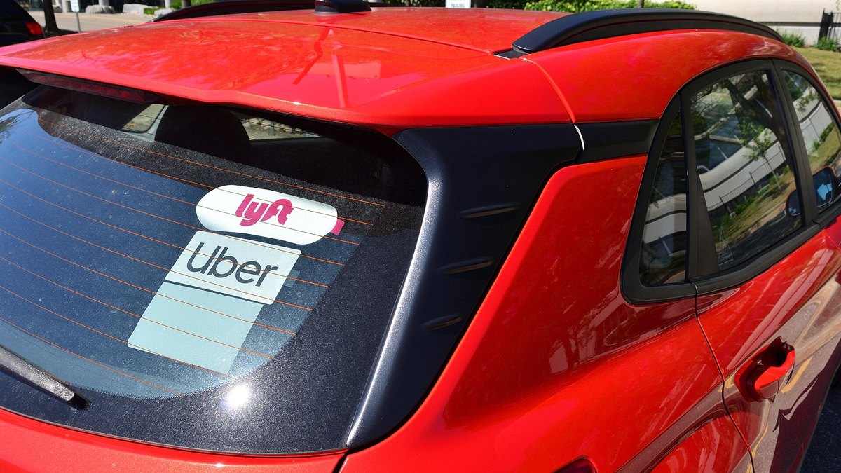 A red car with Lyft and Uber stickers side by side on its rear window, the badge of a driver who works on both rideshare platforms