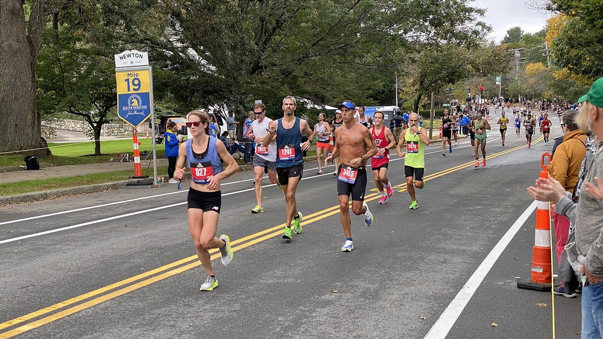 Runners in racing shoes and singlets pass the mile 19 marker on a tree-lined road during the October 2021 Boston Marathon in Newton, Massachusetts