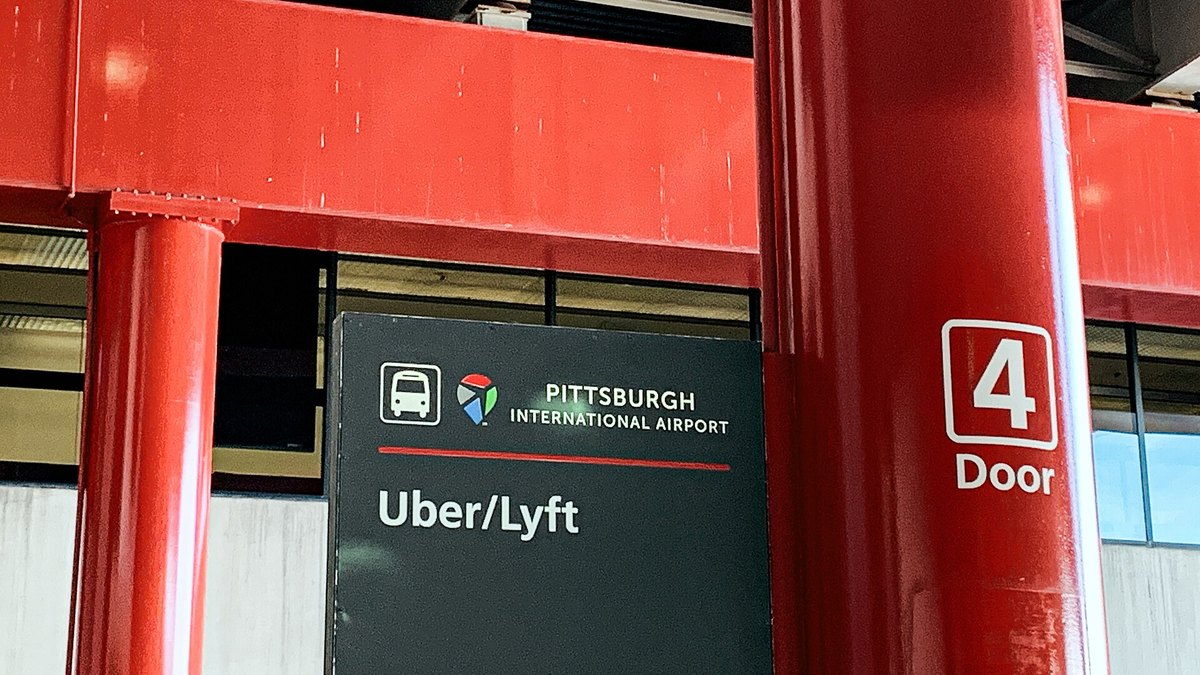 A dark green Uber/Lyft pickup sign beneath red steel beams at door 4 of Pittsburgh International Airport