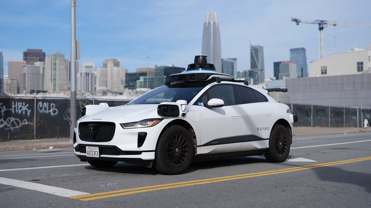 A white Waymo Jaguar I-Pace autonomous taxi with roof sensors drives across a San Francisco street with the downtown skyline behind it