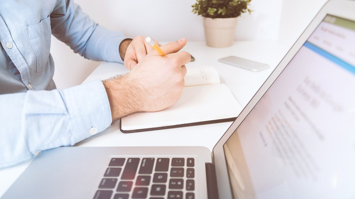 A person's hands resting on a desk beside an open laptop and a notebook with a pencil