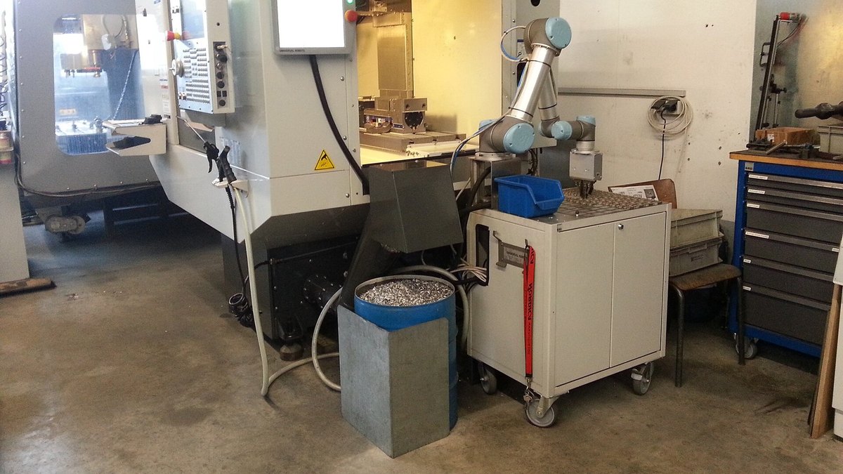A CNC machining center in a machine shop with a collaborative robot arm loading parts and a bin of aluminum chips