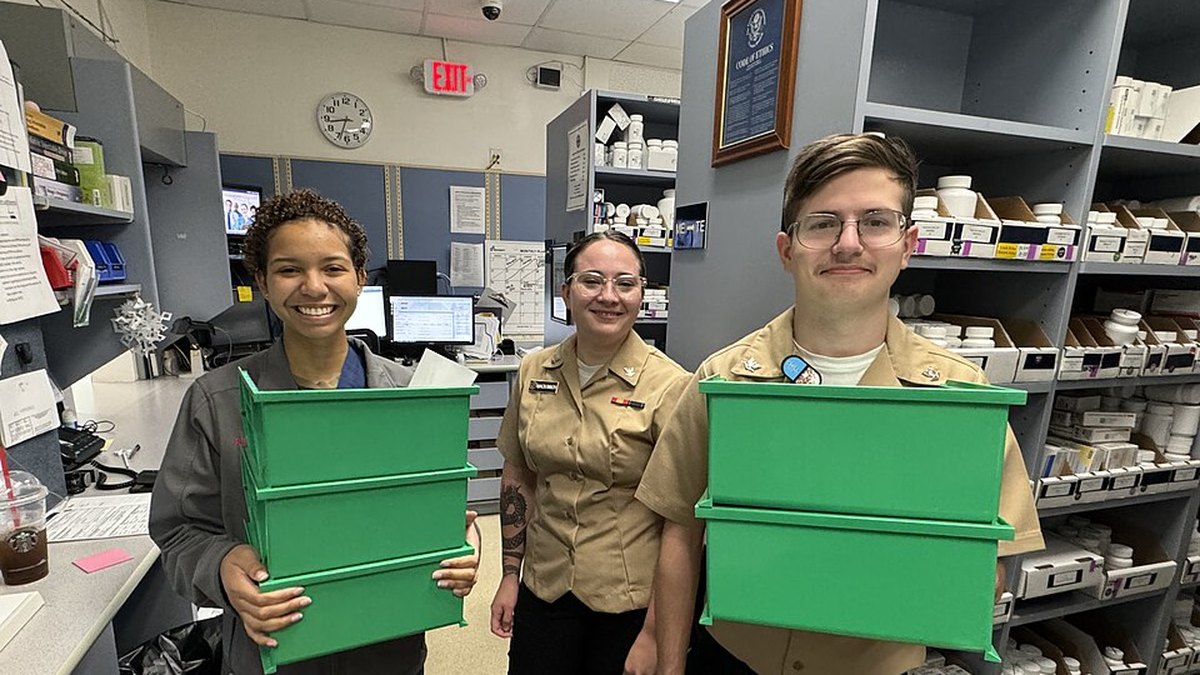 Three pharmacy technicians carrying green medication totes in front of stocked pharmacy shelves