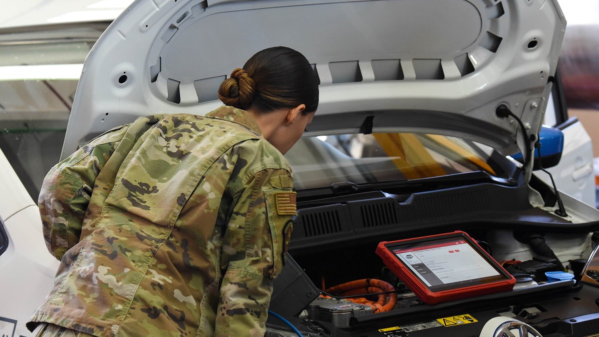 A service member in uniform reading a diagnostic tablet resting on an electric vehicle with its hood open during Air Force EV training