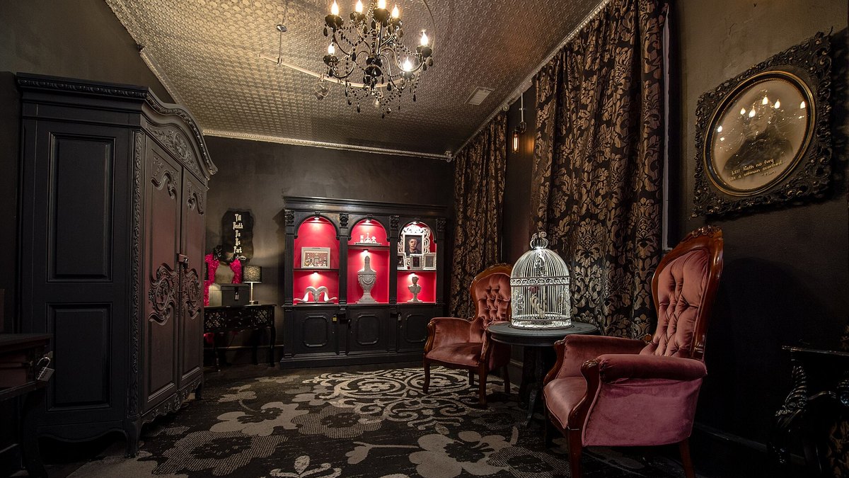 A dark themed escape room set with an armoire, shelves, a birdcage and velvet chairs